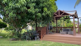 A backyard patio area with a wooden pergola. The pergola has a pitched roof and partially open sides. There are wooden steps leading up to the deck, which features wicker furniture including a chair and two ottomans. The patio is surrounded by lush greenery, including a large tree with dense foliage on the left and various smaller plants. The scene appears tranquil and well-maintained.