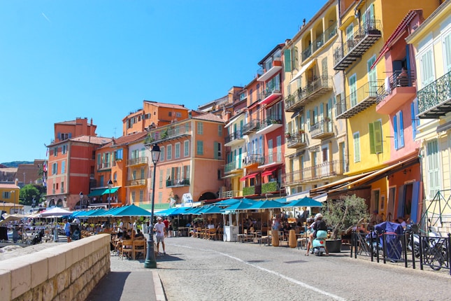 Colorful buildings line a quaint street with outdoor cafes and people enjoying a sunny day. The architecture is vibrant with shades of yellow, orange, red, and pink. Tables and umbrellas are set up outside cafes along the cobblestone street, and a lamp post is visible in the foreground.