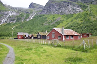A series of rustic, red wooden buildings with white-framed windows and moss-covered roofs situated in a green pastoral area. Mountains with cascading waterfalls form the backdrop, and a dirt path leads up to the houses, which are fenced in with wire and wooden posts.