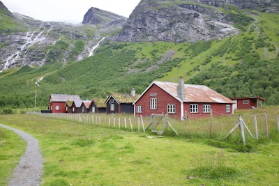 A series of rustic, red wooden buildings with white-framed windows and moss-covered roofs situated in a green pastoral area. Mountains with cascading waterfalls form the backdrop, and a dirt path leads up to the houses, which are fenced in with wire and wooden posts.