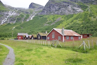 A series of rustic, red wooden buildings with white-framed windows and moss-covered roofs situated in a green pastoral area. Mountains with cascading waterfalls form the backdrop, and a dirt path leads up to the houses, which are fenced in with wire and wooden posts.