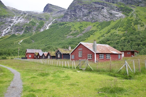 A series of rustic, red wooden buildings with white-framed windows and moss-covered roofs situated in a green pastoral area. Mountains with cascading waterfalls form the backdrop, and a dirt path leads up to the houses, which are fenced in with wire and wooden posts.