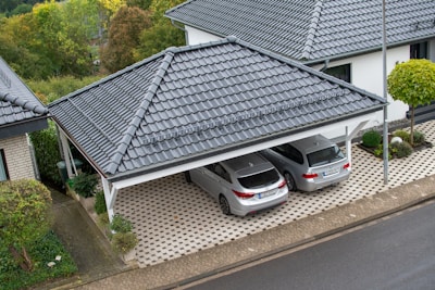 A residential area with two silver cars parked under a covered carport with a tiled roof. The driveway is paved with a patterned design, and there is a well-maintained garden with trees and shrubs nearby. The surrounding area includes other houses and lush greenery indicating a suburban environment.