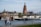 A scenic riverside view features a large cathedral with intricate architecture towering in the background. In front, a classic iron pedestrian bridge spans the river, bustling with people. Below, two boats are moored on the water, one of which displays the name 'Friedrich Rückert'. In the foreground, three people are walking casually along a tree-lined path that follows the riverbank. The scene is framed by leafy green trees, providing a tranquil urban landscape.
