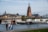 A scenic riverside view features a large cathedral with intricate architecture towering in the background. In front, a classic iron pedestrian bridge spans the river, bustling with people. Below, two boats are moored on the water, one of which displays the name 'Friedrich Rückert'. In the foreground, three people are walking casually along a tree-lined path that follows the riverbank. The scene is framed by leafy green trees, providing a tranquil urban landscape.