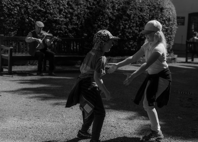 Two children play together in a shaded outdoor area, while a musician plays the violin on a bench nearby. The children appear lively and joyful, and the musician adds a serene, calming presence to the scene.