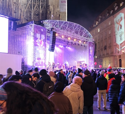A large crowd is gathered in front of a brightly lit outdoor stage at night. The stage is equipped with lighting and sound systems, and a band is performing. The audience is dressed in winter clothing, suggesting a cold season. Some people are holding balloons, and there are buildings in the background with large projections or advertisements on them.