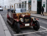 A vintage-style automobile with gold and burgundy accents parked on a street in an urban area. The front of the car features a decorative grill with the word 'Elegant' displayed. The vehicle's design is reminiscent of early 20th-century cars. A figure is walking on the sidewalk beside a building with arched windows and signage.