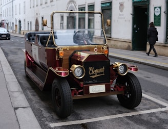 A vintage-style automobile with gold and burgundy accents parked on a street in an urban area. The front of the car features a decorative grill with the word 'Elegant' displayed. The vehicle's design is reminiscent of early 20th-century cars. A figure is walking on the sidewalk beside a building with arched windows and signage.