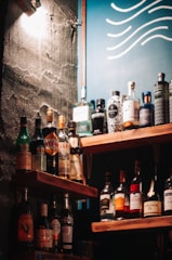 A selection of various liquor bottles is displayed on wooden shelves, set against a wall with a rough texture. A blue board with wavy white lines can be seen in the background, and a warm light casts shadows across the scene.