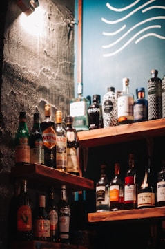 A selection of various liquor bottles is displayed on wooden shelves, set against a wall with a rough texture. A blue board with wavy white lines can be seen in the background, and a warm light casts shadows across the scene.