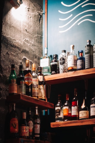 A selection of various liquor bottles is displayed on wooden shelves, set against a wall with a rough texture. A blue board with wavy white lines can be seen in the background, and a warm light casts shadows across the scene.
