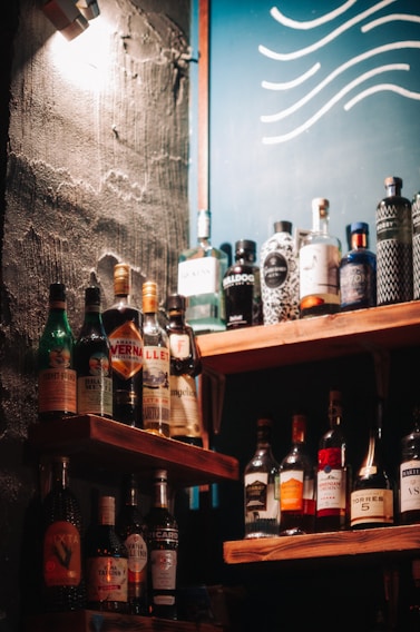 A selection of various liquor bottles is displayed on wooden shelves, set against a wall with a rough texture. A blue board with wavy white lines can be seen in the background, and a warm light casts shadows across the scene.