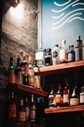 A selection of various liquor bottles is displayed on wooden shelves, set against a wall with a rough texture. A blue board with wavy white lines can be seen in the background, and a warm light casts shadows across the scene.