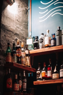 A selection of various liquor bottles is displayed on wooden shelves, set against a wall with a rough texture. A blue board with wavy white lines can be seen in the background, and a warm light casts shadows across the scene.