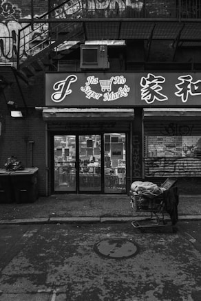 A monochrome image of an urban storefront with signage in both English and Chinese. Graffiti is visible on the building. There is a shopping cart filled with various items on the sidewalk and trash bins to the left. The setting appears gritty and atmospheric with shadows cast by the surrounding structures.