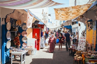 A bustling market scene with narrow streets, offering a variety of goods. Colorful ceramic plates are displayed on a wall, and numerous souvenir items are on tables. People walk through the market, browsing stalls and engaging with vendors. Bright fabrics are draped overhead, providing shade and adding to the vibrant atmosphere.