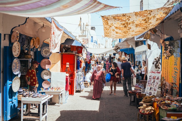 A bustling market scene with narrow streets, offering a variety of goods. Colorful ceramic plates are displayed on a wall, and numerous souvenir items are on tables. People walk through the market, browsing stalls and engaging with vendors. Bright fabrics are draped overhead, providing shade and adding to the vibrant atmosphere.