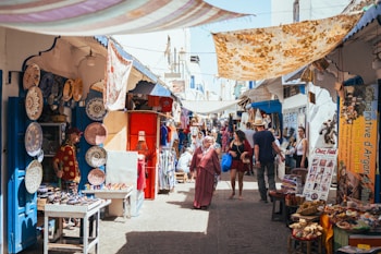 A bustling market scene with narrow streets, offering a variety of goods. Colorful ceramic plates are displayed on a wall, and numerous souvenir items are on tables. People walk through the market, browsing stalls and engaging with vendors. Bright fabrics are draped overhead, providing shade and adding to the vibrant atmosphere.