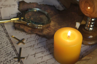 A magnifying glass with a brass frame rests on top of an aged, handwritten document. The composition includes a lit yellow candle casting a gentle glow, adding warmth to the scene. There are also two small brass crosses and parts of a wooden object in the background.
