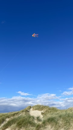 A brightly colored kite with red, orange, and yellow hues flies high in the clear blue sky. Below, there are grassy sand dunes under a sky partially filled with white, fluffy clouds.