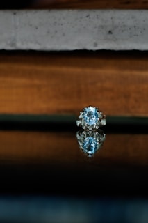 A silver ring with a large blue gemstone is elegantly placed on a reflective surface. The background consists of blurred, textured horizontal layers in shades of grey and brown, emphasizing the focus on the ring.