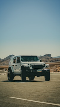 A white off-road vehicle, likely a Jeep Rubicon, is parked in an open area with clear blue skies and mountainous terrain in the background. The vehicle's rugged build and large tires are prominent features. There is minimal vegetation and a road barrier running parallel to the car.