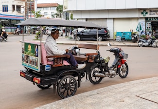 A man is seated in a tuk-tuk, a motorbike with a carriage, parked on a street in an urban area. The vehicle is equipped with a canopy for shade, and the man appears to be holding a phone. Surrounding the tuk-tuk are buildings, other vehicles, and people engaged in daily activities.