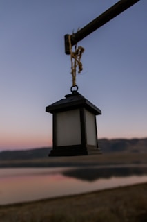 A black lantern hangs from a horizontal metal rod against a backdrop of a serene landscape. The background features a body of water reflecting the soft hues of a twilight sky, with rolling hills silhouetted against the horizon.