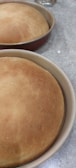 Two freshly baked, golden brown loaves of bread sit in round baking pans on a speckled countertop. The bread is evenly baked, creating a smooth and slightly domed surface.