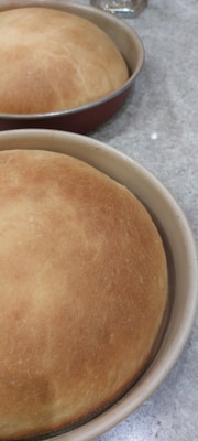 Two freshly baked, golden brown loaves of bread sit in round baking pans on a speckled countertop. The bread is evenly baked, creating a smooth and slightly domed surface.
