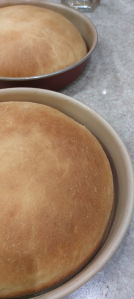 Two freshly baked, golden brown loaves of bread sit in round baking pans on a speckled countertop. The bread is evenly baked, creating a smooth and slightly domed surface.