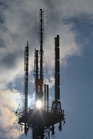 Tall radio towers with multiple antennas positioned on a platform, silhouetted against a vibrant sky with sunlight peeking through the structures. Clouds are scattered in the backdrop, enhancing the dramatic effect of the scene.