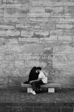 A couple sits closely on a bench against a textured stone wall. Both are leaning in towards each other, conveying intimacy and connection. The setting appears to be urban and minimalist.