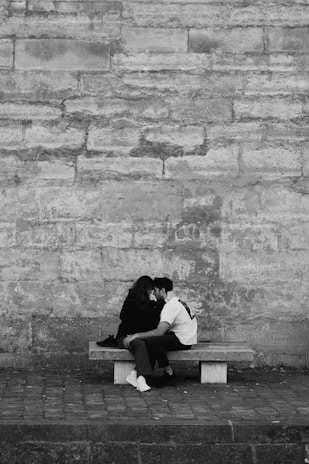 A couple sits closely on a bench against a textured stone wall. Both are leaning in towards each other, conveying intimacy and connection. The setting appears to be urban and minimalist.