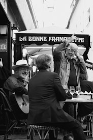 A black and white photograph captures an outdoor café scene. There's a man playing a guitar and smiling warmly. Another man is seated at the table with his back to the camera, engaged in conversation. A third man stands with an expressive gesture, looking cheerful. The setting includes a canopy with the words 'La Bonne Franquette', and several wine glasses are placed on the table.