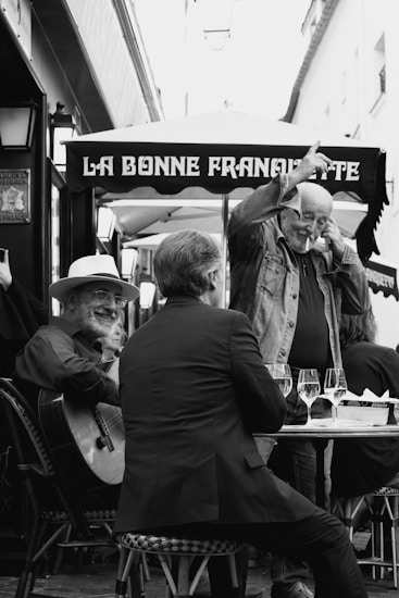 A black and white photograph captures an outdoor caf&eacute; scene. There's a man playing a guitar and smiling warmly. Another man is seated at the table with his back to the camera, engaged in conversation. A third man stands with an expressive gesture, looking cheerful. The setting includes a canopy with the words 'La Bonne Franquette', and several wine glasses are placed on the table.