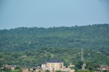A large house with a dark roof is situated at the base of a lush, green forested hill. Several other smaller buildings and structures are visible nearby, as well as a tall power line tower. The image conveys a sense of tranquility and natural beauty, with the dense greenery of the forest dominating the scene.