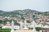 A panoramic view of a cityscape with a mixture of residential buildings spread across a hilly area. In the foreground, there are several tall, white structures with green domes, resembling minarets. The background is dominated by a lush green hill, and the sky is overcast.