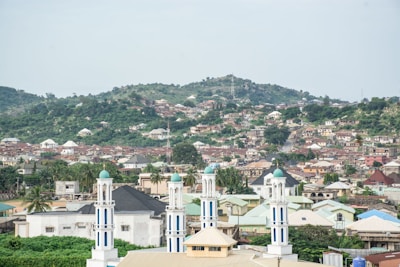 A panoramic view of a cityscape with a mixture of residential buildings spread across a hilly area. In the foreground, there are several tall, white structures with green domes, resembling minarets. The background is dominated by a lush green hill, and the sky is overcast.