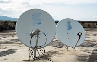 Two satellite dishes are positioned on a flat, weathered rooftop. The dishes are white with a blue logo, and the background shows a cloudy sky and distant mountains.