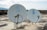 Two satellite dishes are positioned on a flat, weathered rooftop. The dishes are white with a blue logo, and the background shows a cloudy sky and distant mountains.