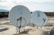 Two satellite dishes are positioned on a flat, weathered rooftop. The dishes are white with a blue logo, and the background shows a cloudy sky and distant mountains.