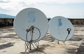 Two satellite dishes are positioned on a flat, weathered rooftop. The dishes are white with a blue logo, and the background shows a cloudy sky and distant mountains.