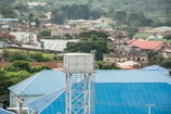 A water tank is elevated above a large blue-roofed building, amidst a hilly suburban area. Various residential structures with different colored roofs are visible, surrounded by lush green vegetation and scattered trees. The setting suggests a serene residential region with ample greenery.