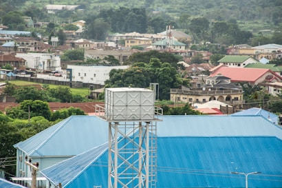 A water tank is elevated above a large blue-roofed building, amidst a hilly suburban area. Various residential structures with different colored roofs are visible, surrounded by lush green vegetation and scattered trees. The setting suggests a serene residential region with ample greenery.