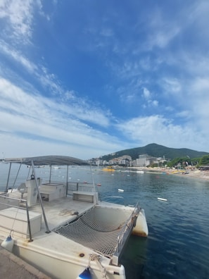 A catamaran docked by a calm waterfront, with a distant view of modern buildings nestled against lush green hills under a blue sky with scattered clouds. The area is vibrant with various colored buoys in the water and a sandy beach nearby.