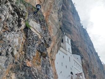A white building with a tower is nestled against a massive rocky cliff. The structure appears to be a religious site, possibly a monastery or church, and is integrated into the natural landscape. A black lamp is mounted on the rock face, adding an element of human presence to the rugged environment.
