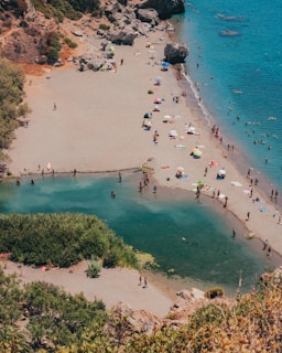 A serene beach scene featuring a sandy shore with numerous people enjoying leisure activities. Colorful umbrellas dot the beach, providing shade to visitors. The clear blue sea contrasts with the verdant greenery surrounding a small lagoon. Some rocks and vegetation are visible on the left side, creating a natural border.