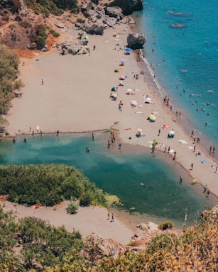A serene beach scene featuring a sandy shore with numerous people enjoying leisure activities. Colorful umbrellas dot the beach, providing shade to visitors. The clear blue sea contrasts with the verdant greenery surrounding a small lagoon. Some rocks and vegetation are visible on the left side, creating a natural border.
